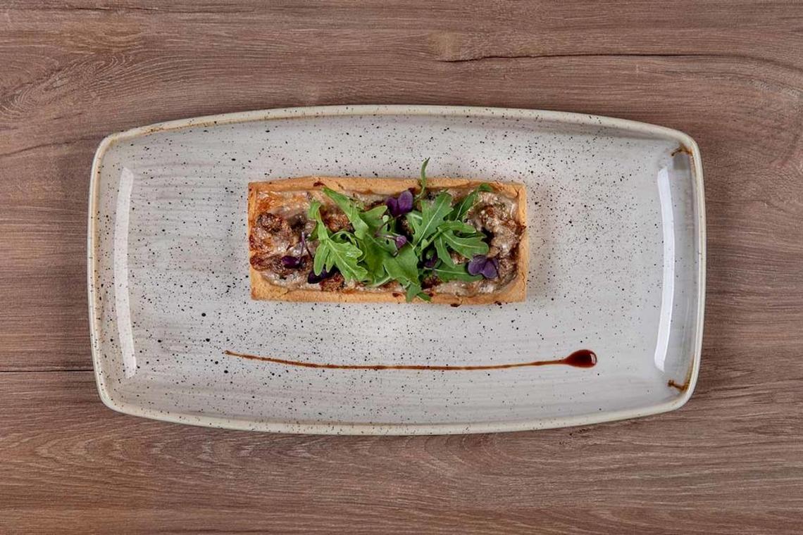 Gebackenes Brot mit Fleisch und Salat auf einem weißen Teller mit schwarzen Punkten auf einem Holztisch.