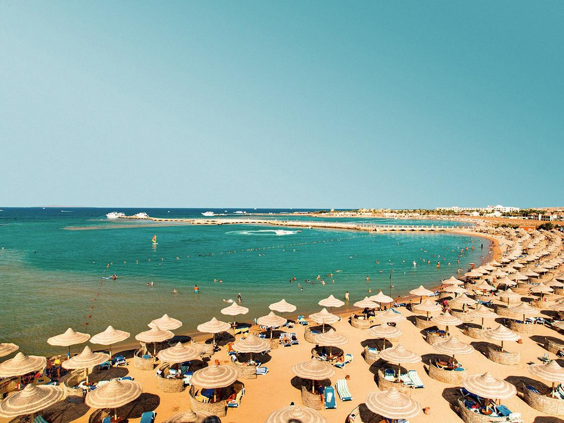 Luftaufnahme des Strandes des Sentido Mamlouk Palace Hotels mit vielen Sonnenschirmen und Gästen im Wasser.