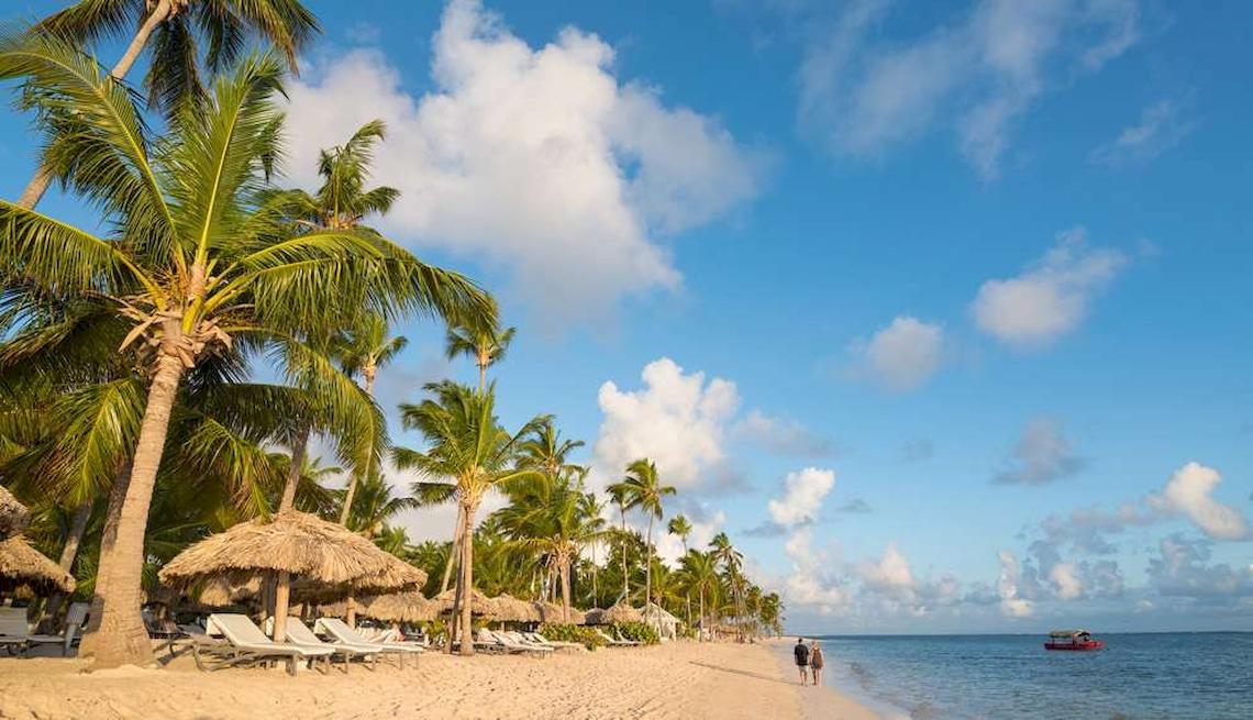 Strand des Catalonia Royal Bavaro mit Palmen, Sonnenliegen und zwei Personen, die am Wasser entlanggehen