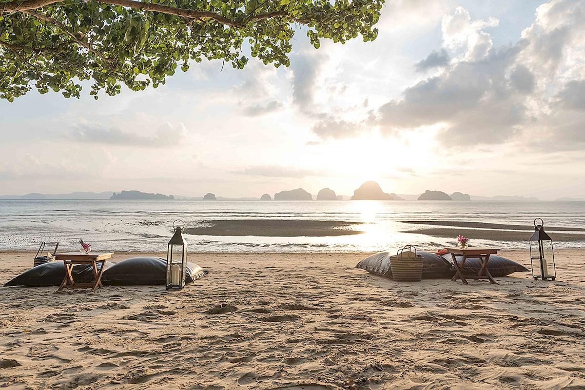 Strand des Tubkaak Krabi Boutique Resorts bei Sonnenuntergang mit Tischen, Kissen und Laternen.