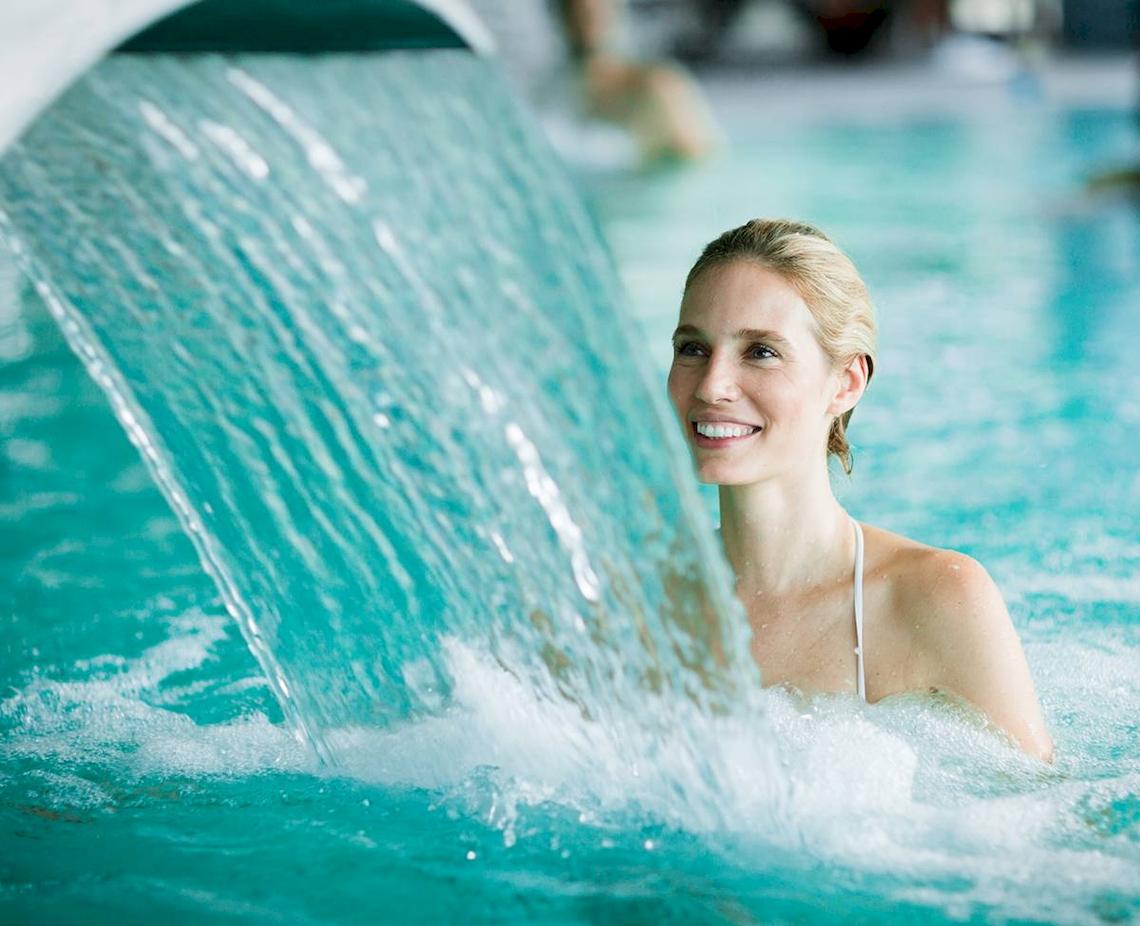 Frau im Schwimmbad unter einem Wasserfall, lächelt ins Bild