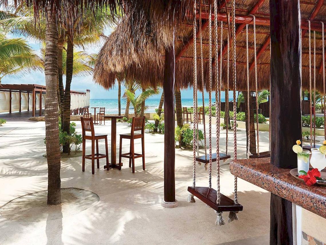 Strandbar des El Dorado Royale a Spa Resort by Karisma mit Tischen, Stühlen und einer Schaukel am Strand.