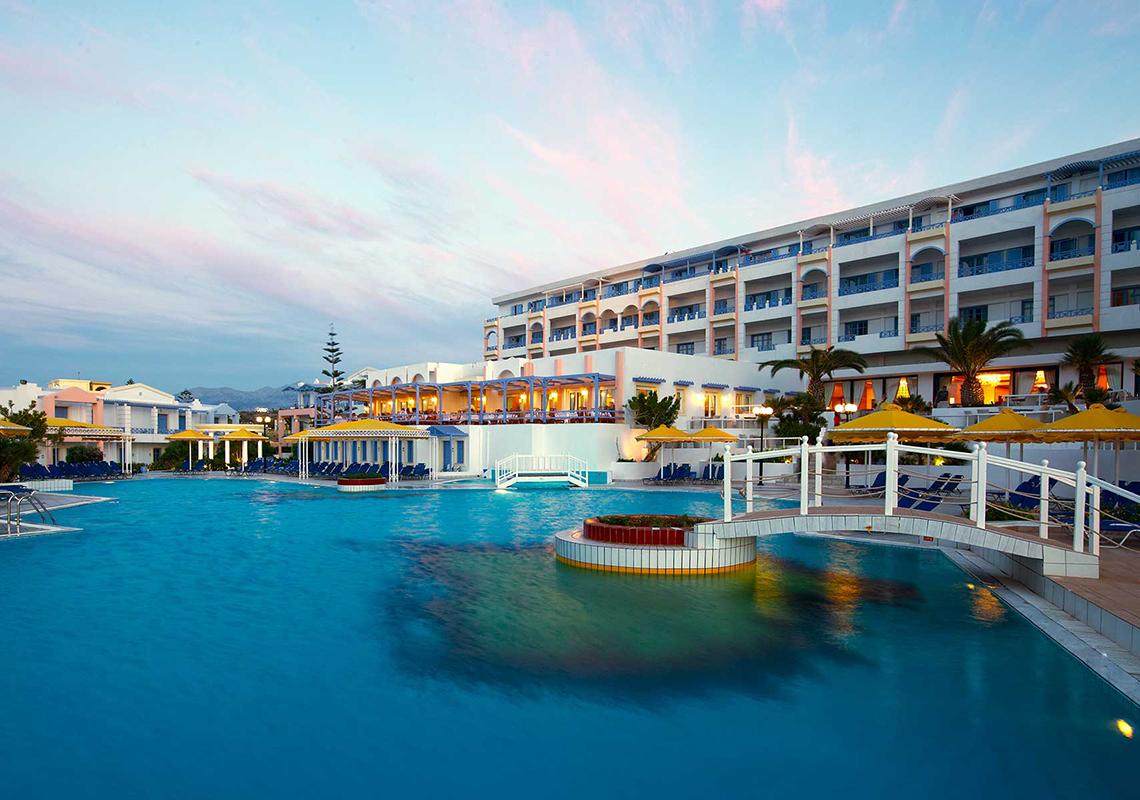 Außenpool des Serita Beach Hotels mit Brücke und Sonnenschirmen bei Sonnenuntergang