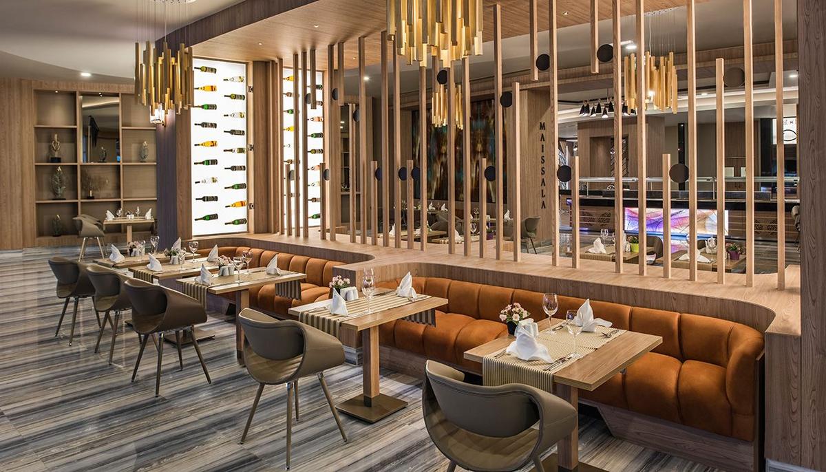 Innenansicht des Restaurants im Delphin BE Grand Resort mit braunen Sofas, Holztischen und goldenen Kronleuchtern.