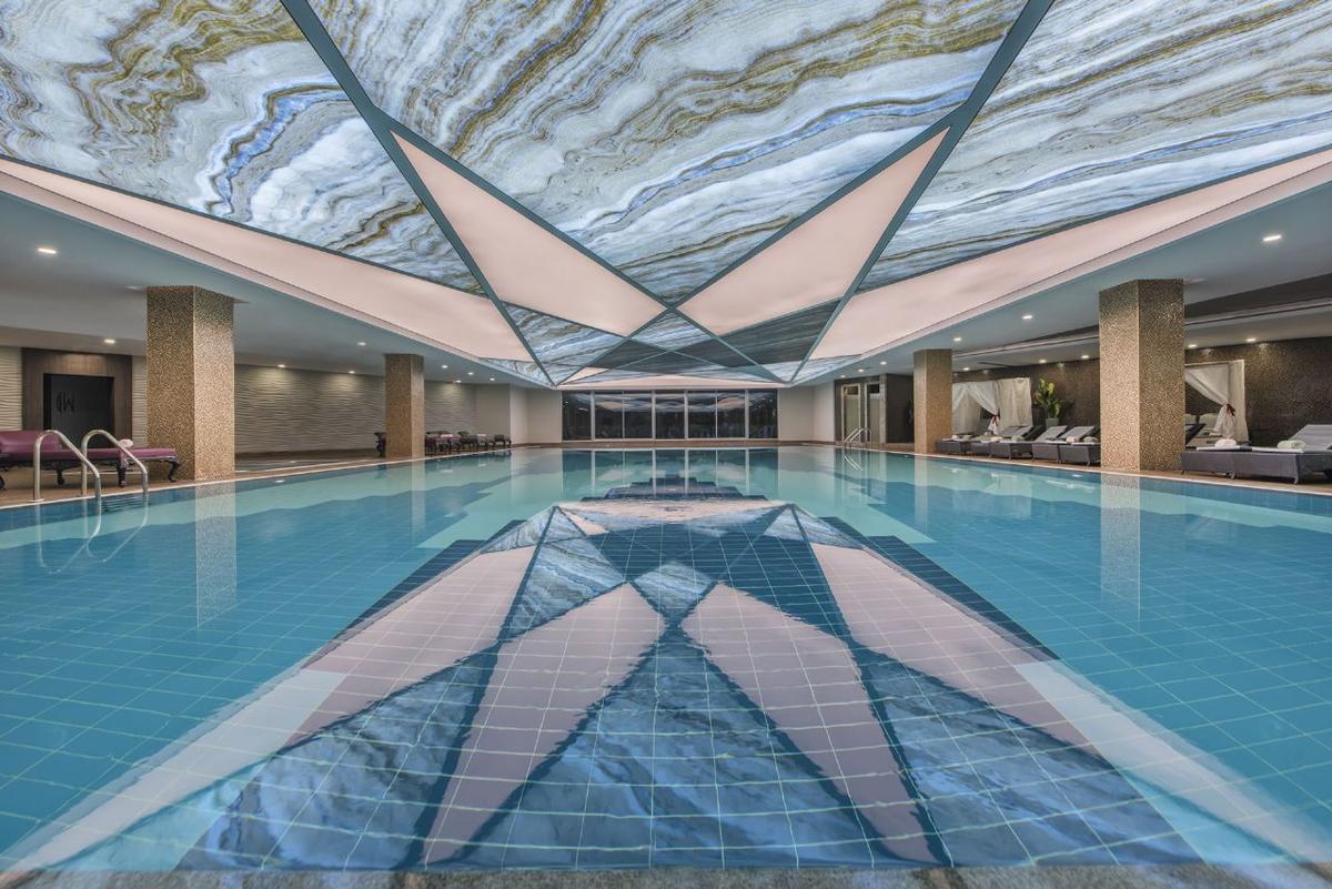 Innenpool des Delphin BE Grand Resort mit geometrischer Decke und blauem Wasser.