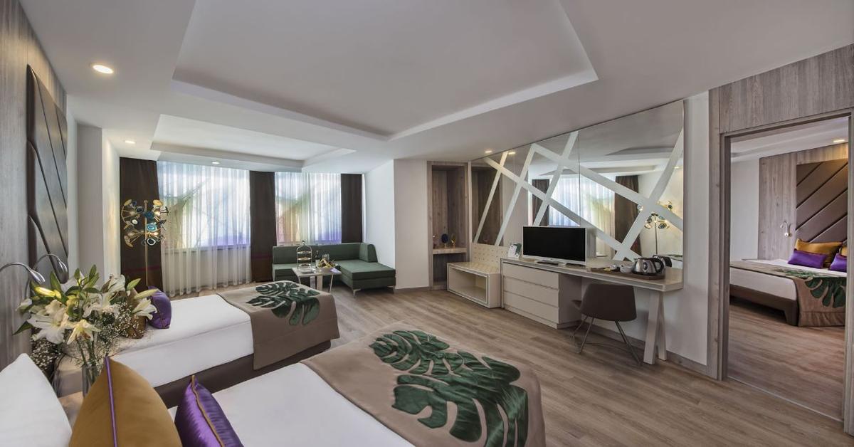 Innenansicht eines Doppelzimmers im Delphin BE Grand Resort mit zwei Betten, einem Schreibtisch und einem Fernseher.