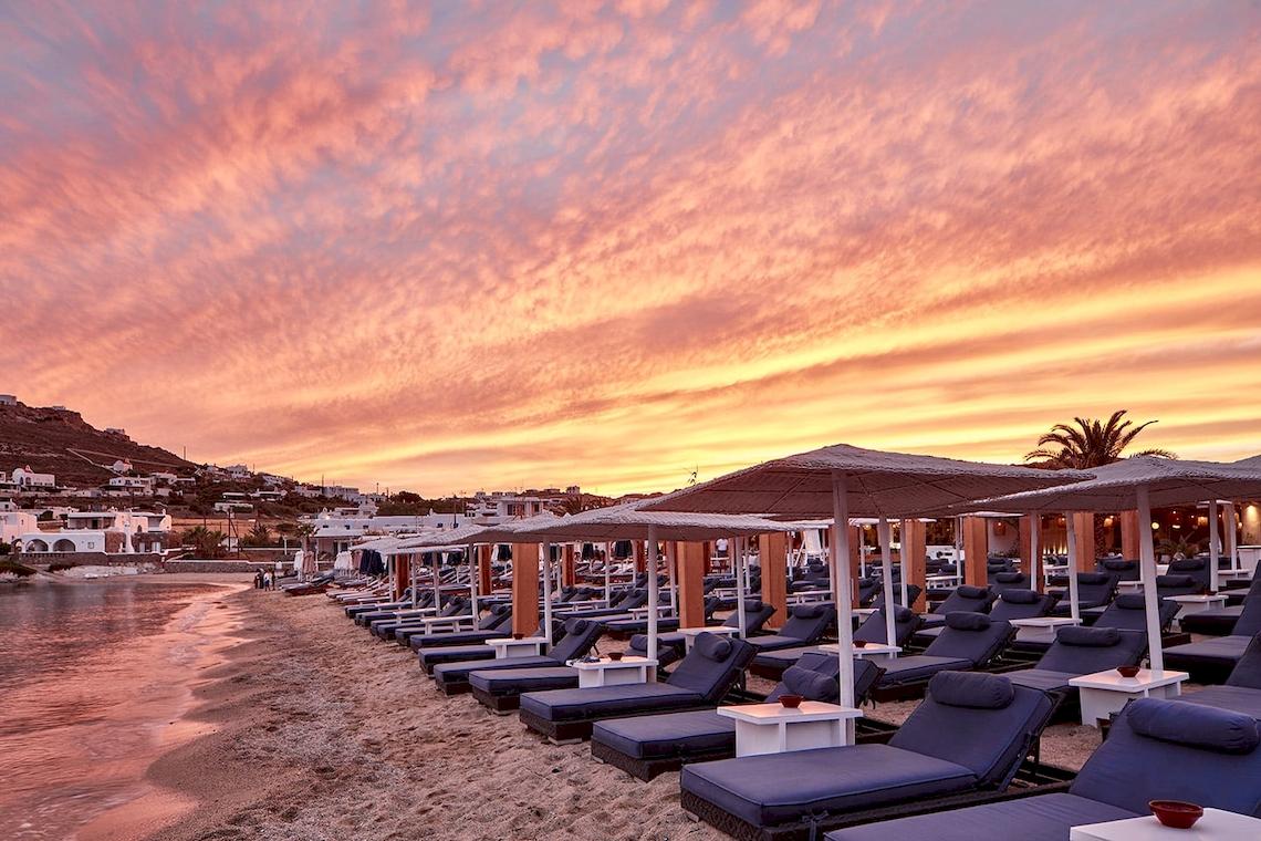 Strand des Mykonos Ammos Hotels mit vielen Liegen und Sonnenschirmen bei Sonnenuntergang