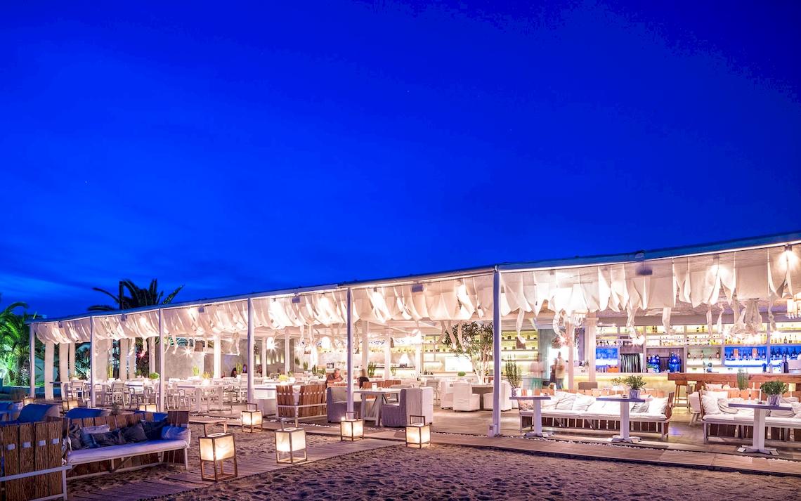 Nachtansicht des Mykonos Ammos Hotel-Strand-Lounge-Bereichs mit weißen Möbeln und Beleuchtung.