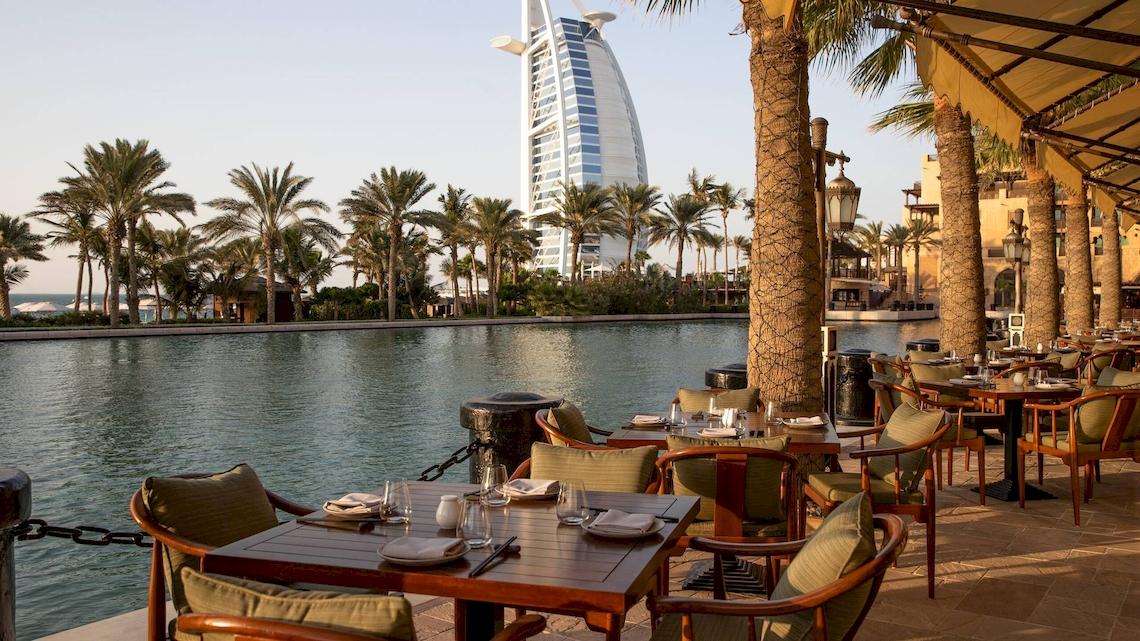 Außenbereich des Jumeirah Mina A'Salam Hotels mit einem Restaurant am Wasser, Blick auf die Palmen und das Burj Al Arab Hotel.