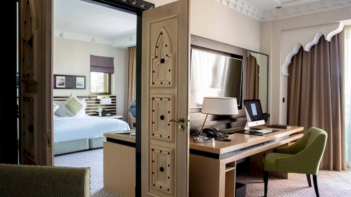 Ein Hotelzimmer mit Schreibtisch, grünem Stuhl und Laptop, Bett und Fenstern.