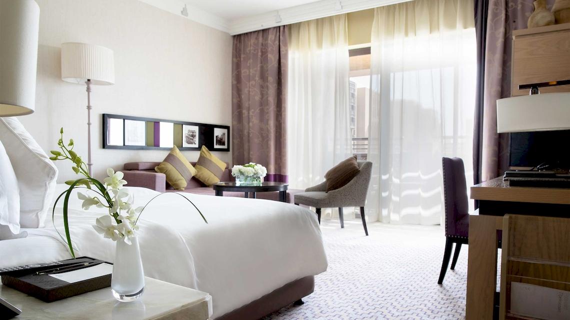 Innenansicht eines Hotelzimmers im Jumeirah Mina A'Salam mit Bett, Tisch, Stuhl und Fenstern.