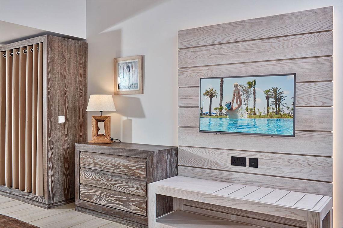 Inneneinrichtung eines Zimmers im Mare Ayia Napa Hotel mit Fernseher und Holzmöbeln.