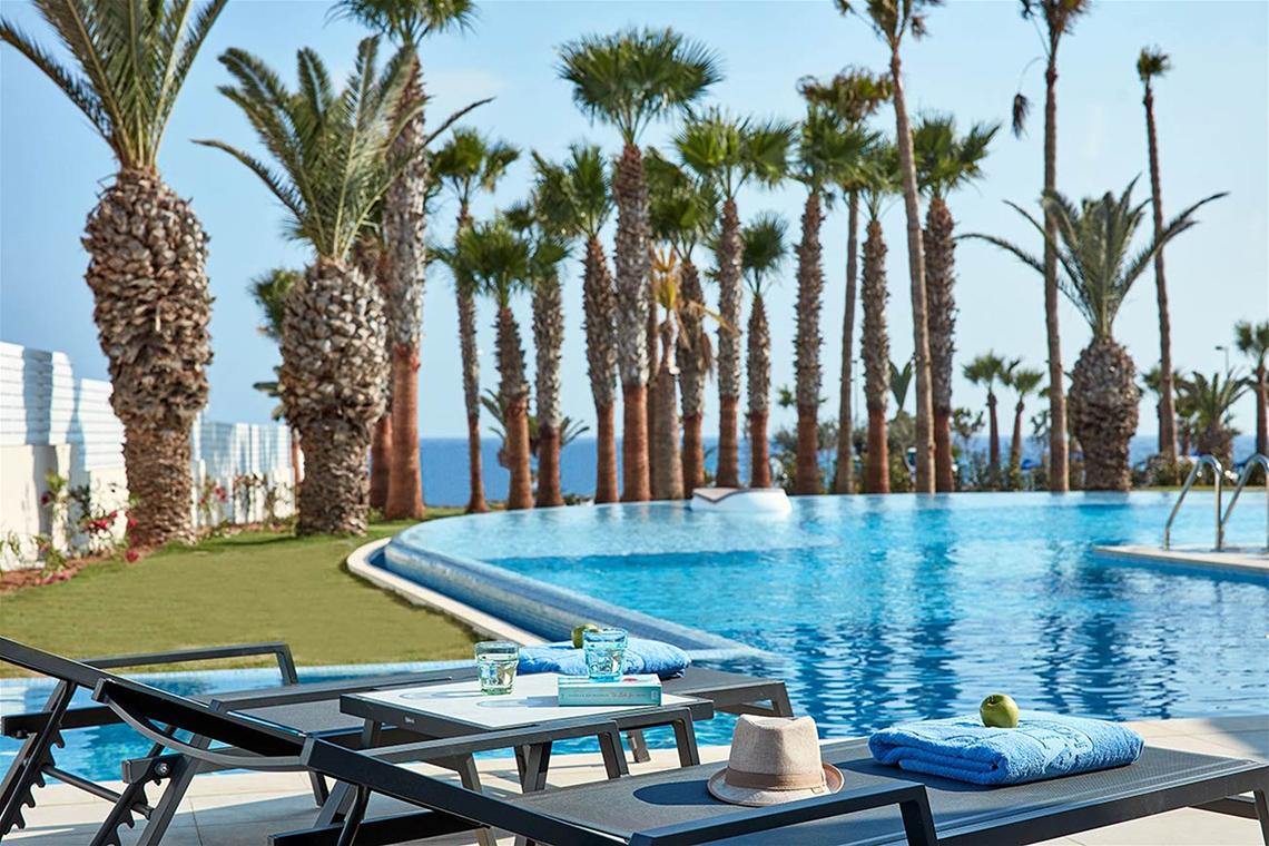 Außenpool des Mare Ayia Napa Hotels mit Sonnenliegen, Handtüchern und Blick auf das Meer.