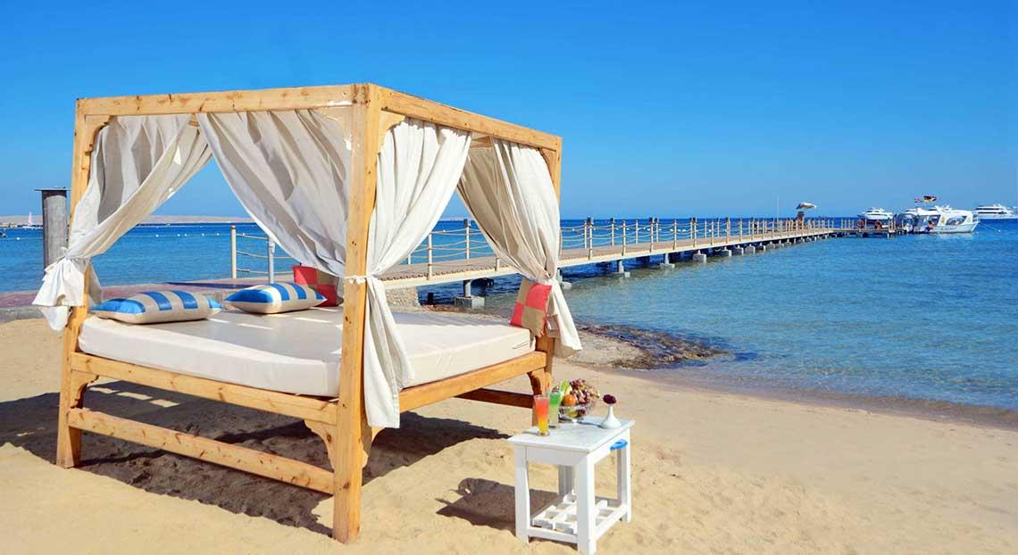 Ein Bett am Strand des Pickalbatros White Beach Resorts mit einem weißen Vorhang und einer kleinen Tisch daneben.