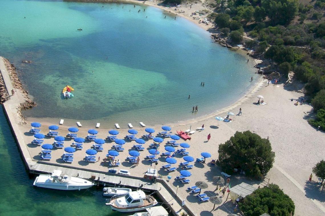 Luftaufnahme des Hotel Ollastu Resorts mit Strand, blauen Sonnenschirmen und Booten im Wasser.