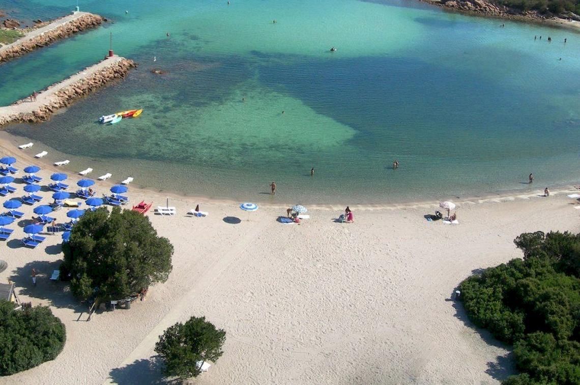 Luftaufnahme des Hotel Ollastu Resorts mit weißer Strand, blauen Sonnenschirmen und Menschen im Wasser.
