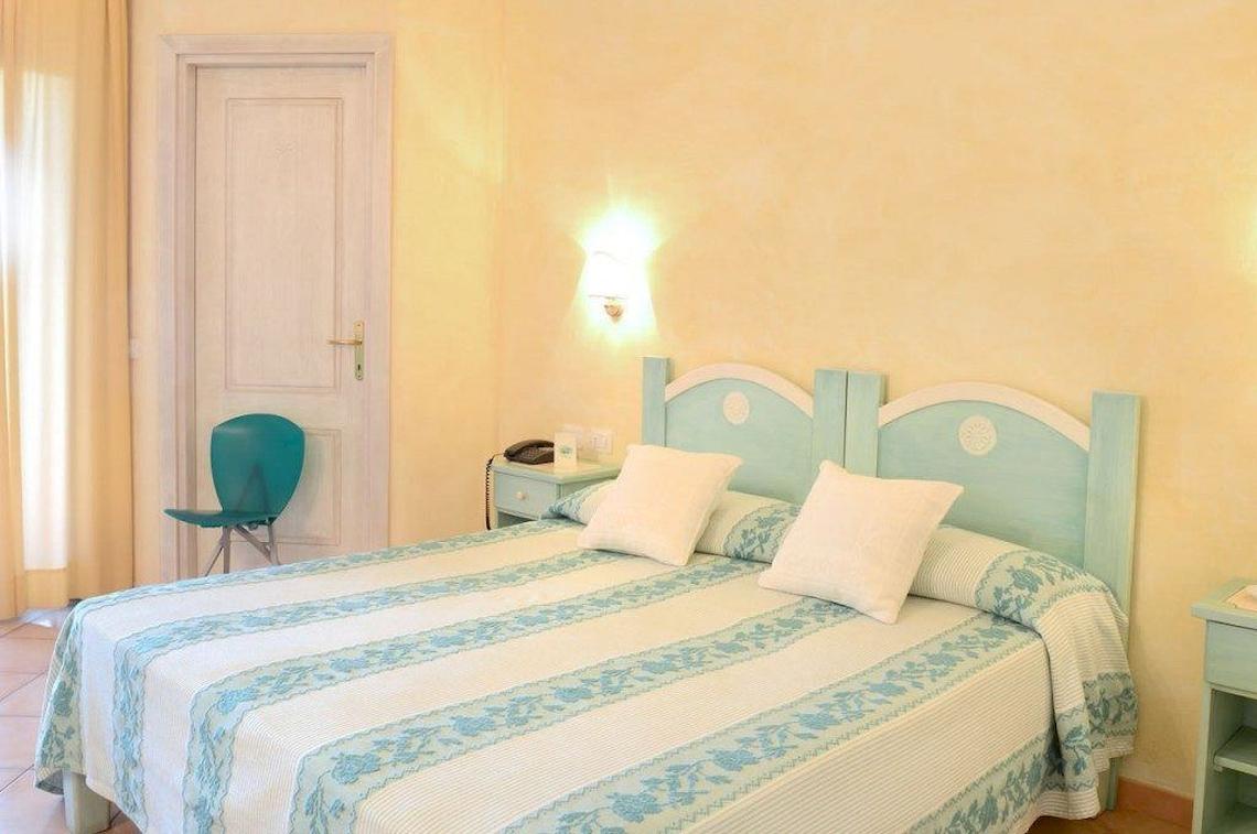 Doppelbettzimmer im Hotel Ollastu Resort mit blauem Kopfteil und gelben Wänden.