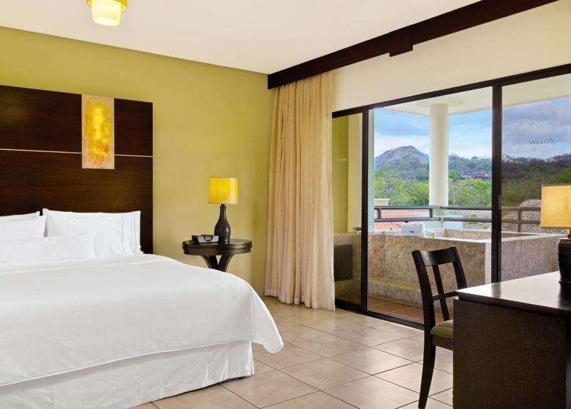 Innenansicht eines Hotelzimmers im Westin Reserva Conchal, mit Bett, Tisch, Lampen und Balkon.