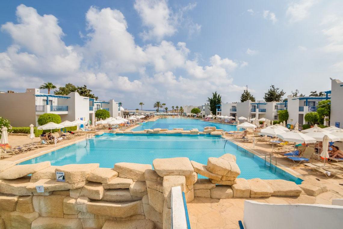 Außenpool im Callisto Holiday Village mit umliegenden Liegestühlen und Gebäuden.