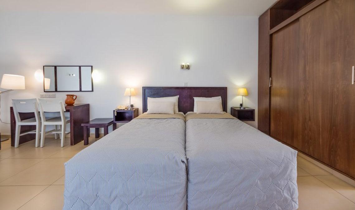 Innenansicht eines Doppelzimmers im Callisto Holiday Village mit zwei Betten, Nachttischen und einer Lampe.