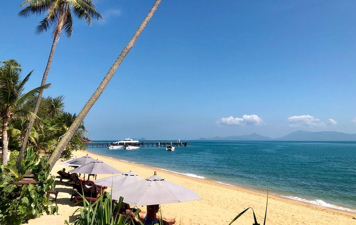 Strandansicht des Saree Samui Resorts mit Palmen, Sonnenschirmen und einer Brücke im Hintergrund.
