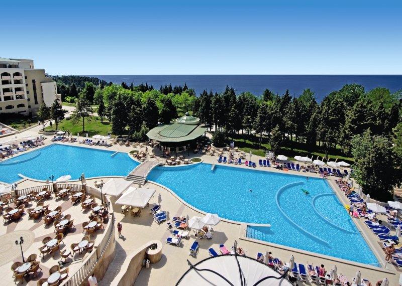 Luftaufnahme des Poolbereichs im Sol Nessebar Palace All Inclusive Hotel mit zwei Schwimmbecken, Sonnenliegen, Bäumen und Meer im Hintergrund.