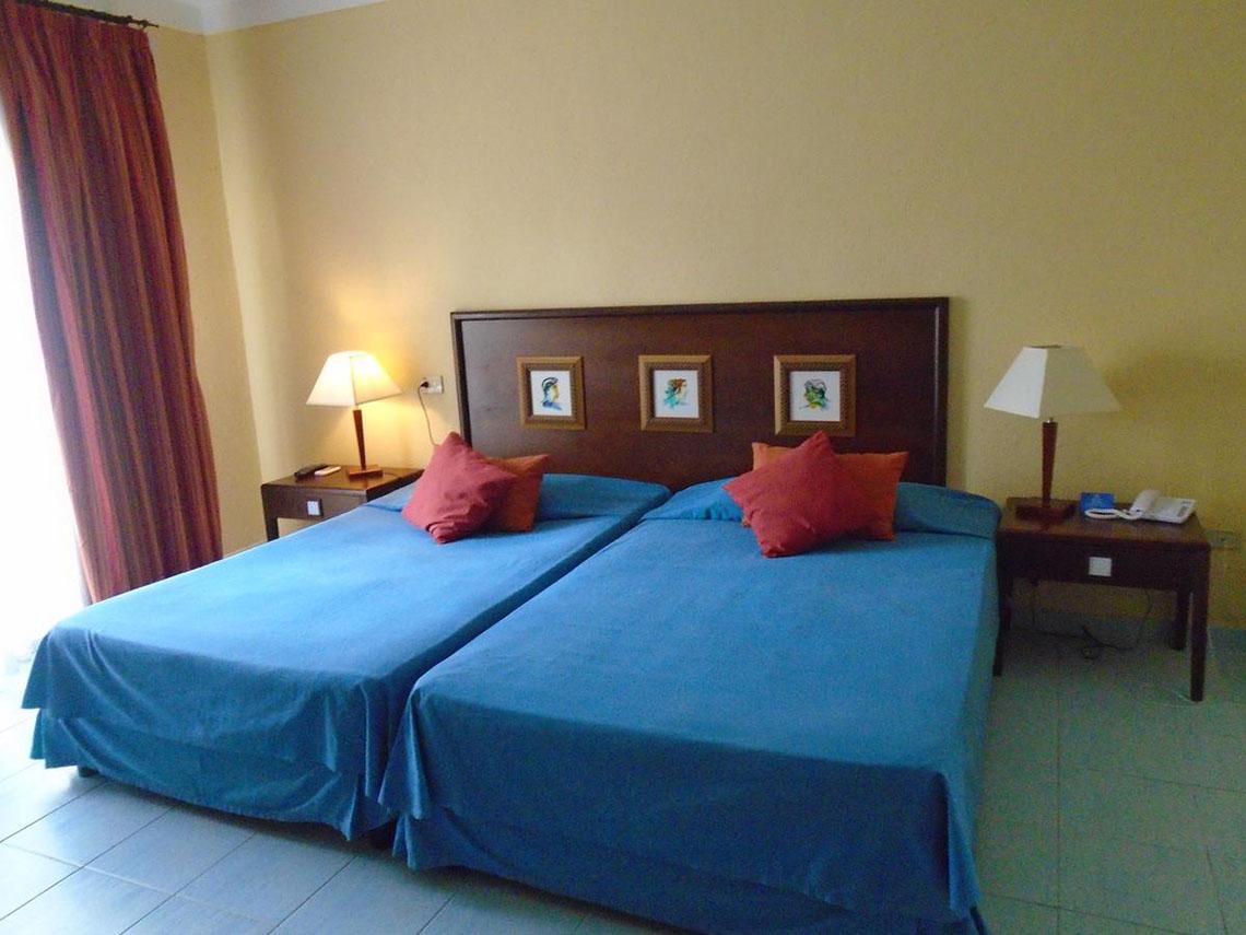 Doppelbett-Zimmer im ROC Arenas Doradas Hotel mit gelben Wänden, blauen Laken und roten Kissen.