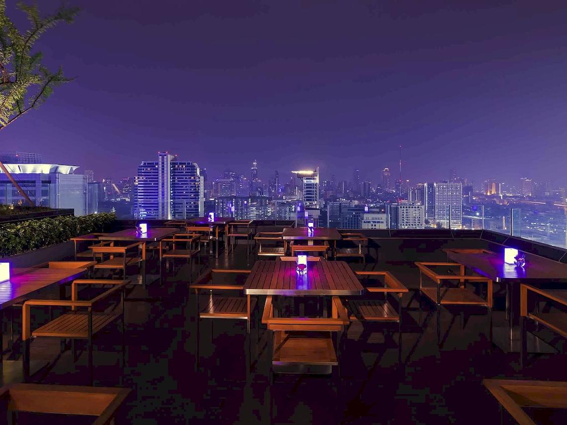Nachtansicht der Dachterrasse des Mercure Bangkok Siam Hotels mit blau beleuchteten Tischen und Stühlen.