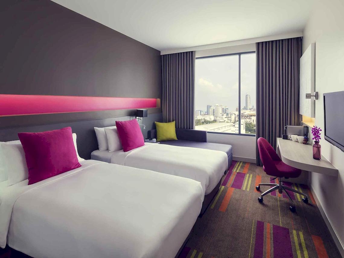 Innenansicht eines Doppelzimmers im Mercure Bangkok Siam Hotel mit zwei Betten, Sofa und Schreibtisch.