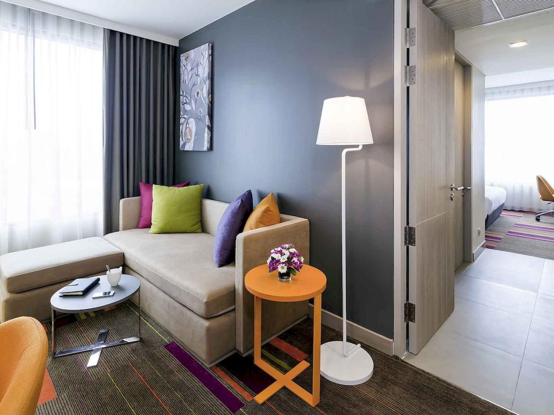 Innenansicht eines Hotelzimmers im Mercure Bangkok Siam mit einer gemütlichen Sitzecke, einem Tisch mit Blumen und einer Stehlampe.