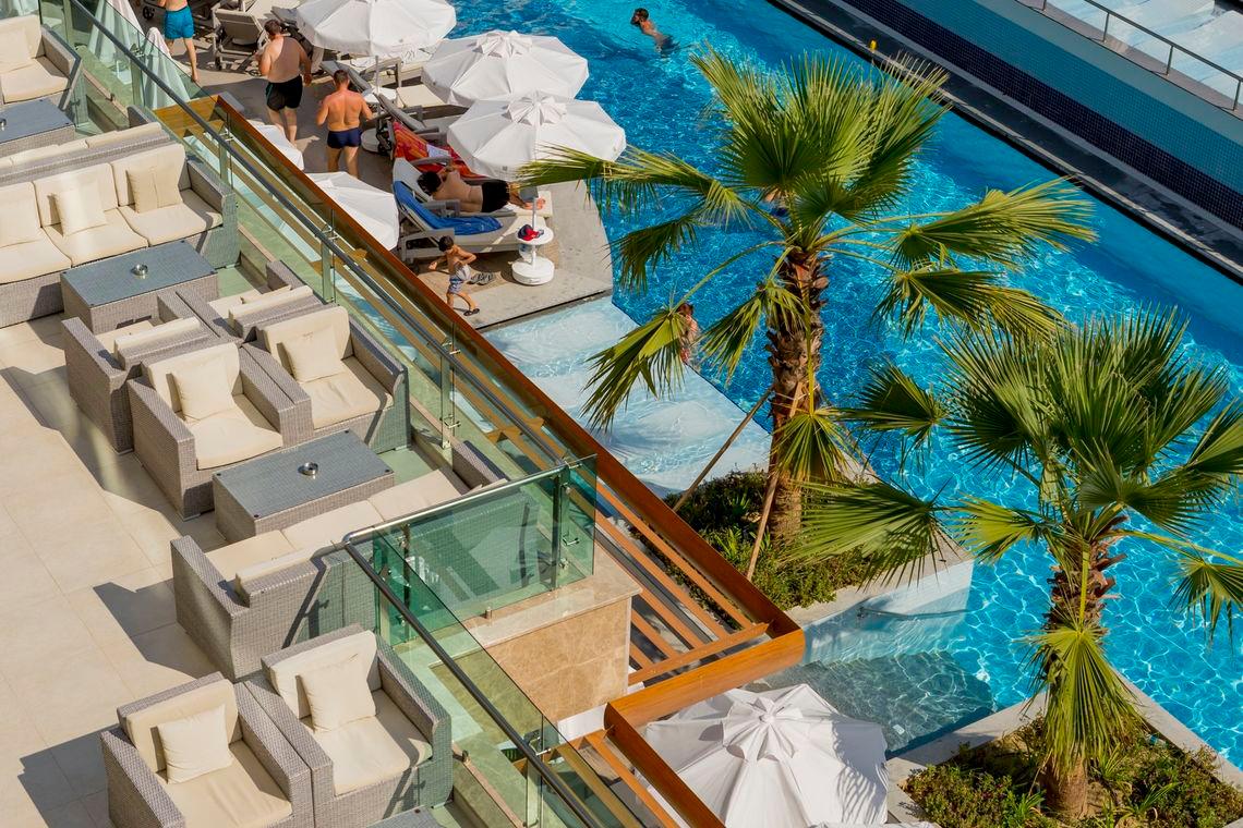 Ein Hotelpoolbereich mit Liegestühlen, Sonnenschirmen und Palmen, wo einige Gäste schwimmen und entspannen.