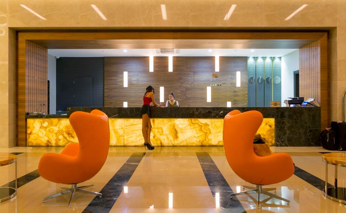 Empfangsbereich des Side Sungate Hotels mit Rezeption und orangefarbenen Stühlen.