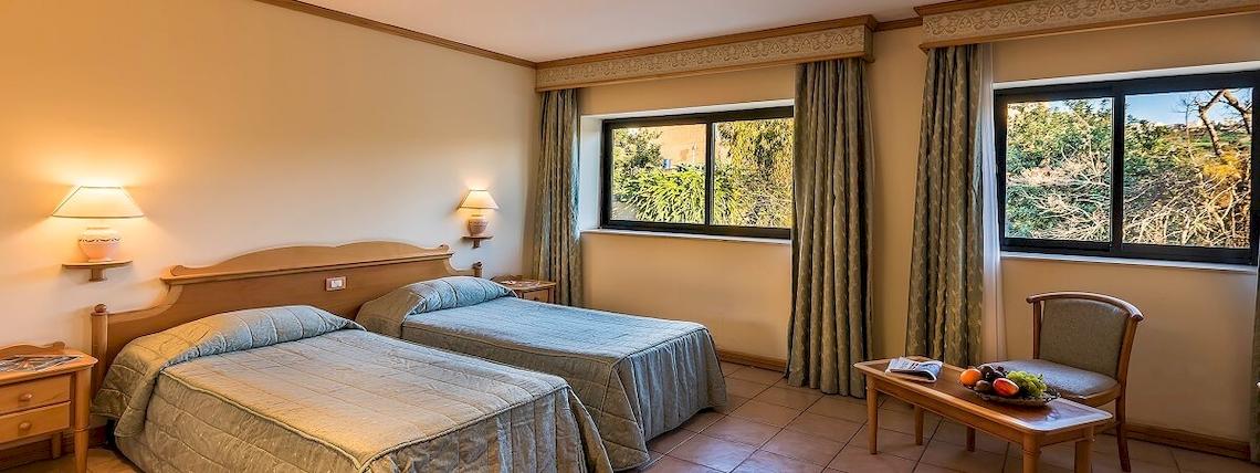Doppelzimmer im Grand Hotel Gozo mit zwei Betten, Fenstern, Vorhängen, Lampen und einem Tisch mit Obst.