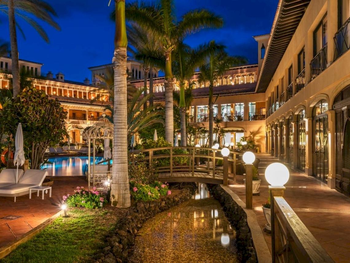 Abendansicht des Secrets Bahia Real Resort & Spa mit beleuchtetem Gehweg, Brücke und Swimmingpool.