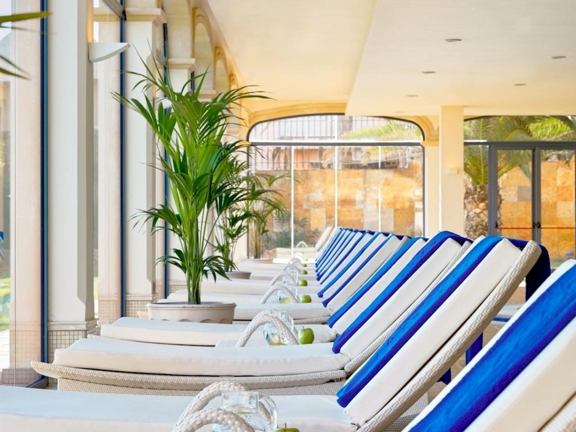 Reihe von weißen und blauen Sonnenliegen im Innenbereich des Secrets Bahia Real Resort & Spa mit Pflanzen und Glasfenstern im Hintergrund.