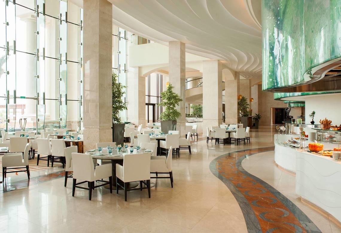 Innenbereich des St. Regis Saadiyat Island Resorts mit weißen Tischen und Stühlen, Glaswänden und Pflanzen.