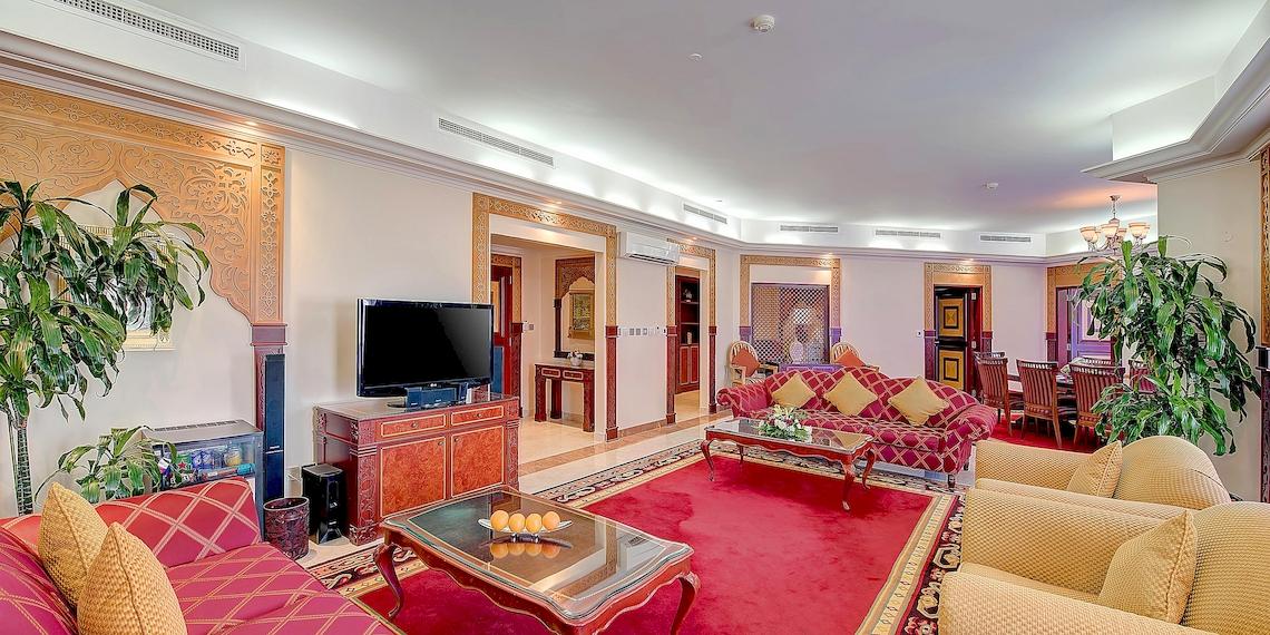 Wohnzimmer im Crowne Plaza Resort Salalah mit rotem Teppich, roten Sofas, Fernseher, Tisch und Pflanzen.