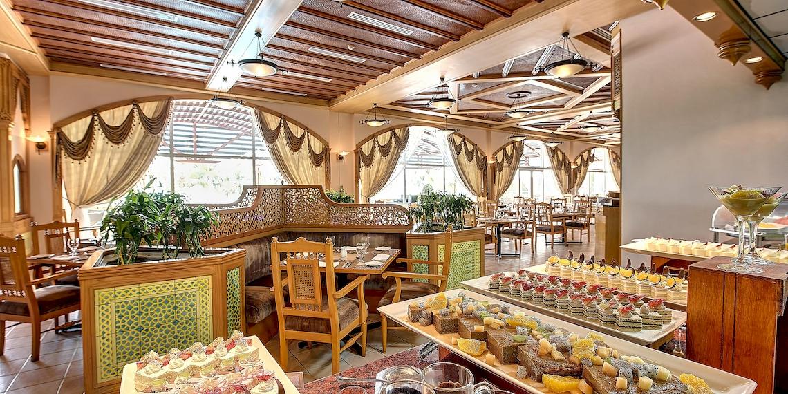 Innenansicht des Restaurants im Crowne Plaza Resort Salalah mit Holzmöbeln und einer Auswahl an Desserts.