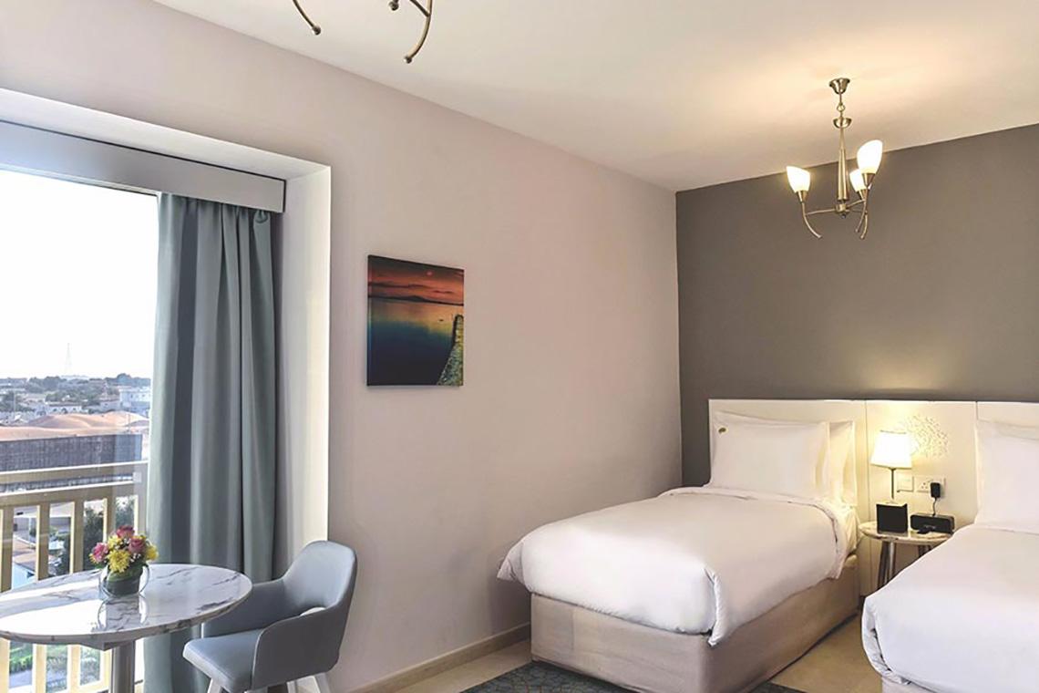 Innenansicht eines Hotelzimmers von Jannah Hotels Resorts & Villas mit zwei Betten, einem Tisch und einem Balkon.
