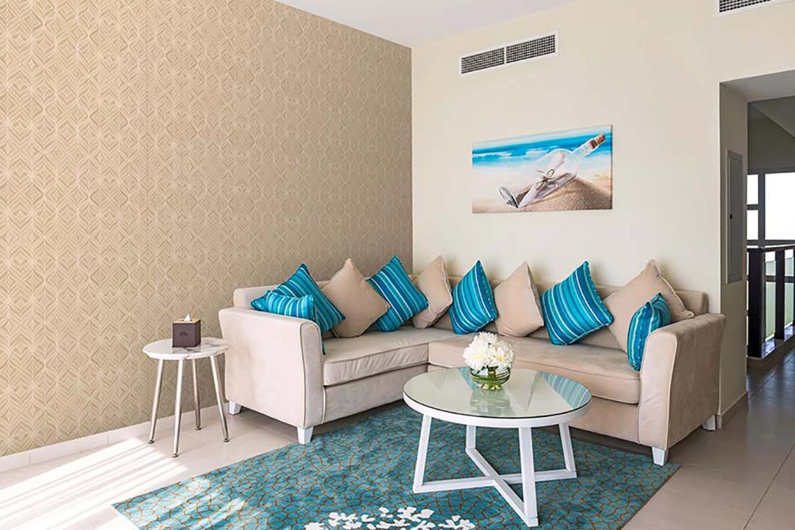 Wohnzimmer mit beigem Sofa, blauen Kissen, Tisch mit Blumenvase und dekorativem Bild an der Wand.