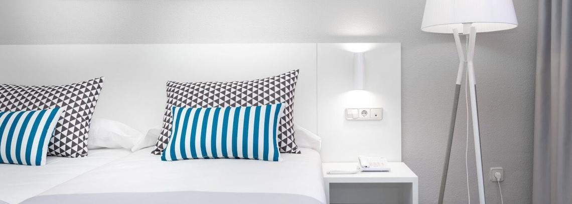 Ein modernes Hotelzimmer mit einem Bett, weißen Kopfteilen, drei Kissen, einer Lampe und einer Steckdose.