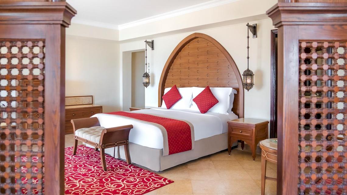 Innenansicht eines eleganten Hotelzimmers im Kempinski Hotel Soma Bay mit einem großen Bett, roten Kissen und zwei Nachttischen.