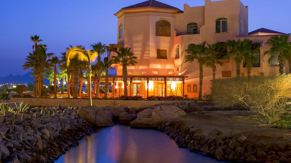 Das Kempinski Hotel Soma Bay ist bei Nacht mit warmem Licht beleuchtet, umgeben von Palmen und Felsen.