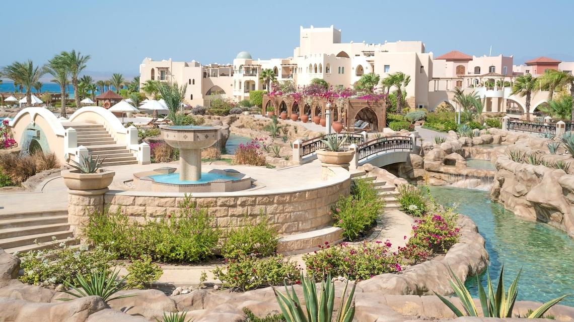 Außenansicht des Kempinski Hotels Soma Bay mit einem Brunnen, einer Brücke und tropischen Pflanzen.