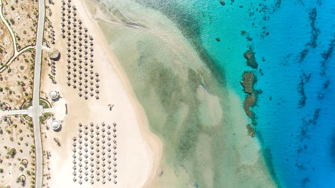 Luftaufnahme des Kempinski Hotels Soma Bay am Strand mit vielen Sonnenschirmen und blauem Wasser.