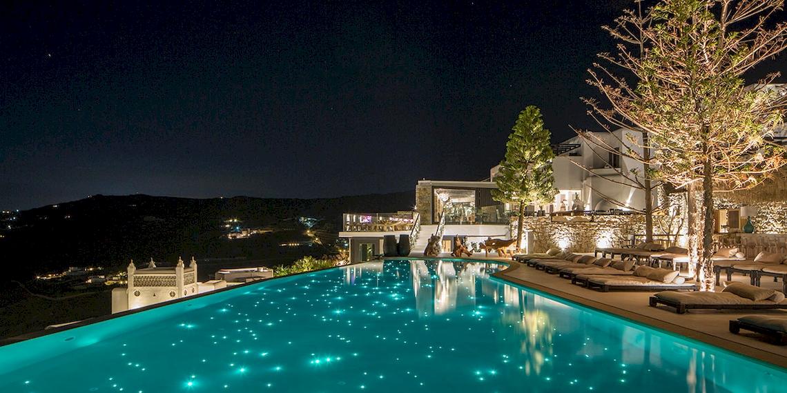 Nachtansicht des Pools im Myconian Utopia Resort mit beleuchteten Bäumen und Loungeliegen.