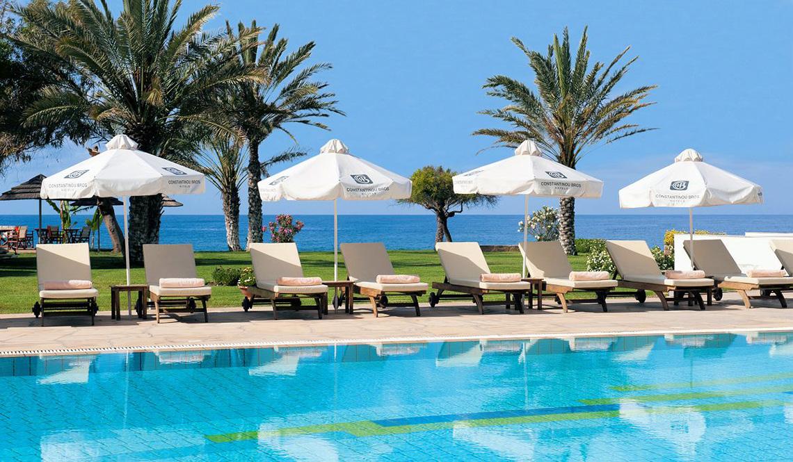 Reihe von Sonnenliegen und Sonnenschirmen am Pool des Athena Royal Beach Hotel mit Blick auf das Meer.