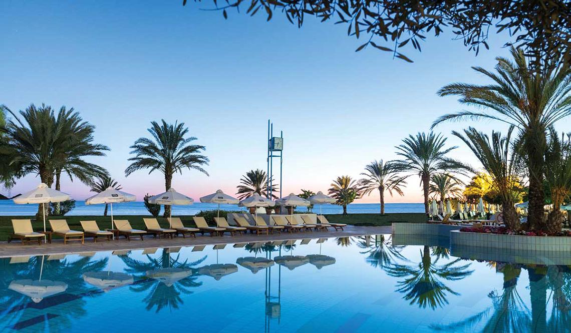 Poolbereich des Athena Royal Beach Hotel bei Abendstimmung mit Palmen und Sonnenliegen