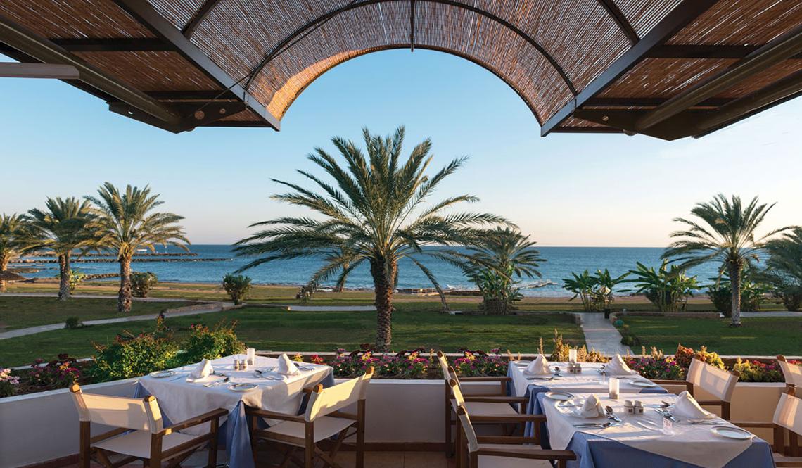 Außenterrasse des Athena Royal Beach Hotel mit Blick auf das Meer und Palmen.
