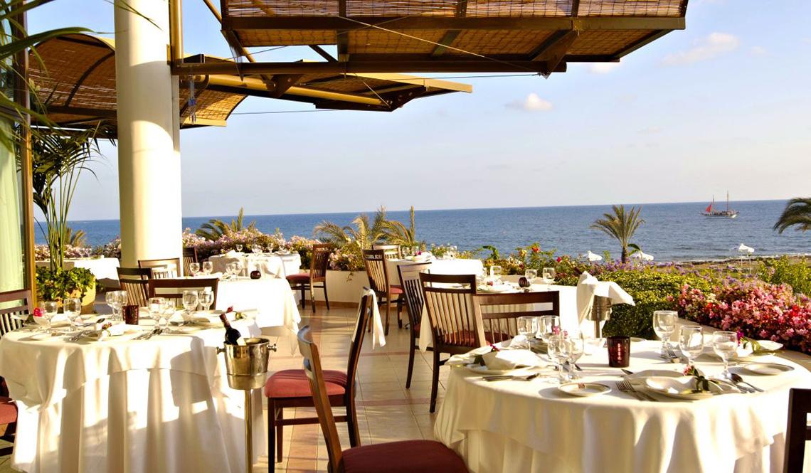 Außenbereich des Athena Royal Beach Hotels mit Tisch und Stühlen, Blick auf das Meer.